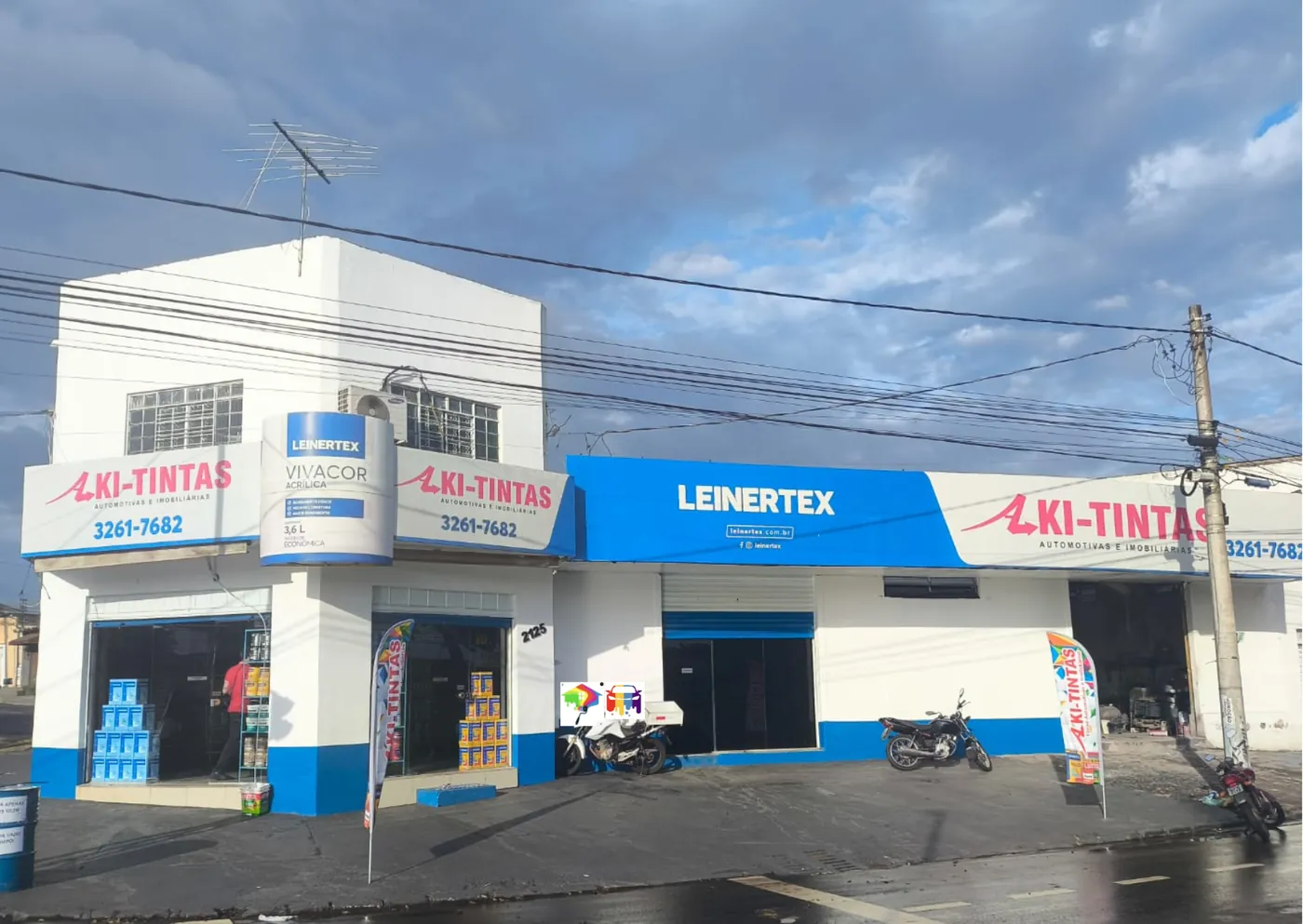 Fachada da loja AKI-TINTAS, com placas da marca, vitrines com latas de tinta empilhadas, letreiro azul da Leinertex e bandeiras de divulgação na calçada